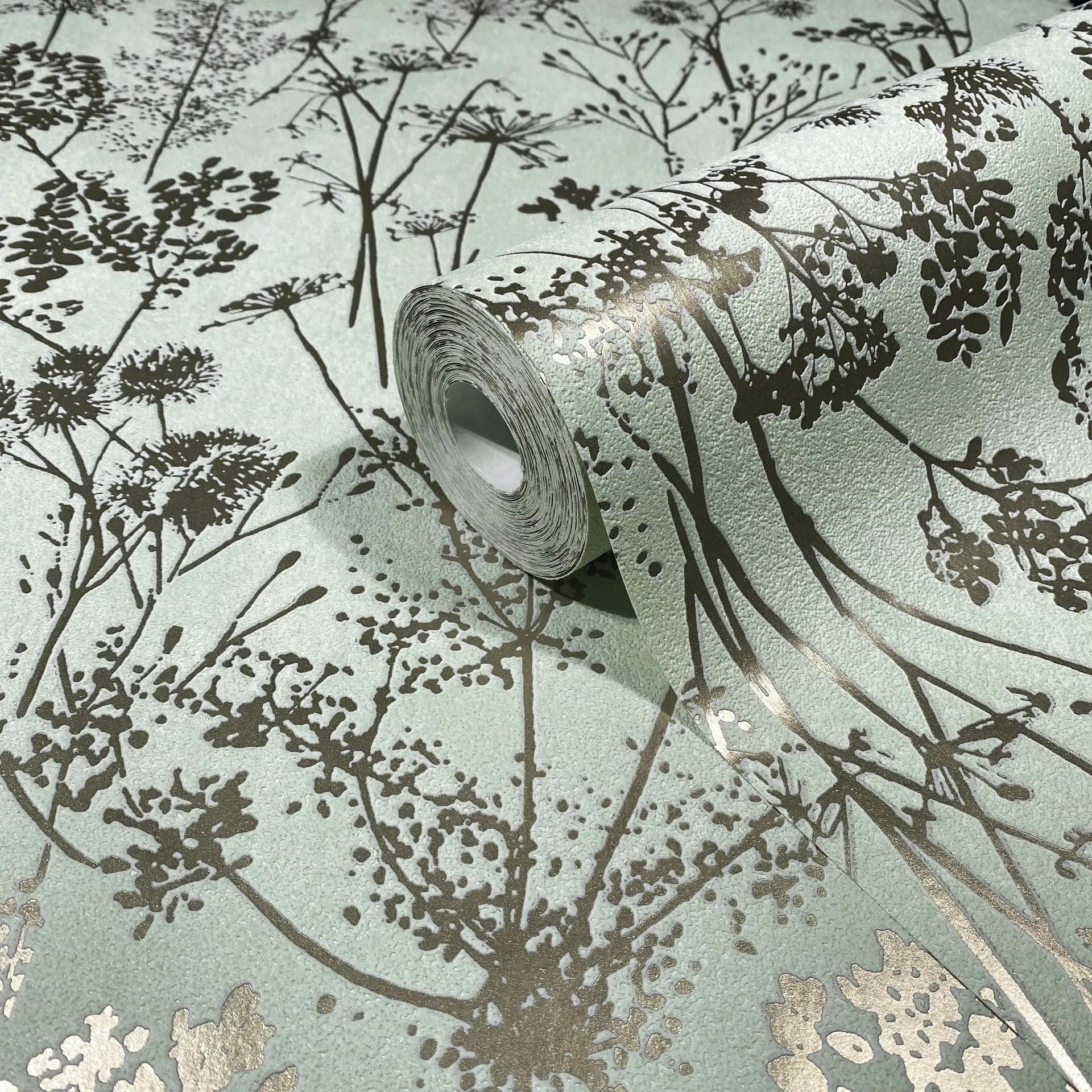 Marburg Mona 82465 Close-up of a wallpaper roll showing metallic gold botanical silhouettes on a pale green textured background, with the pattern unrolled beneath it.