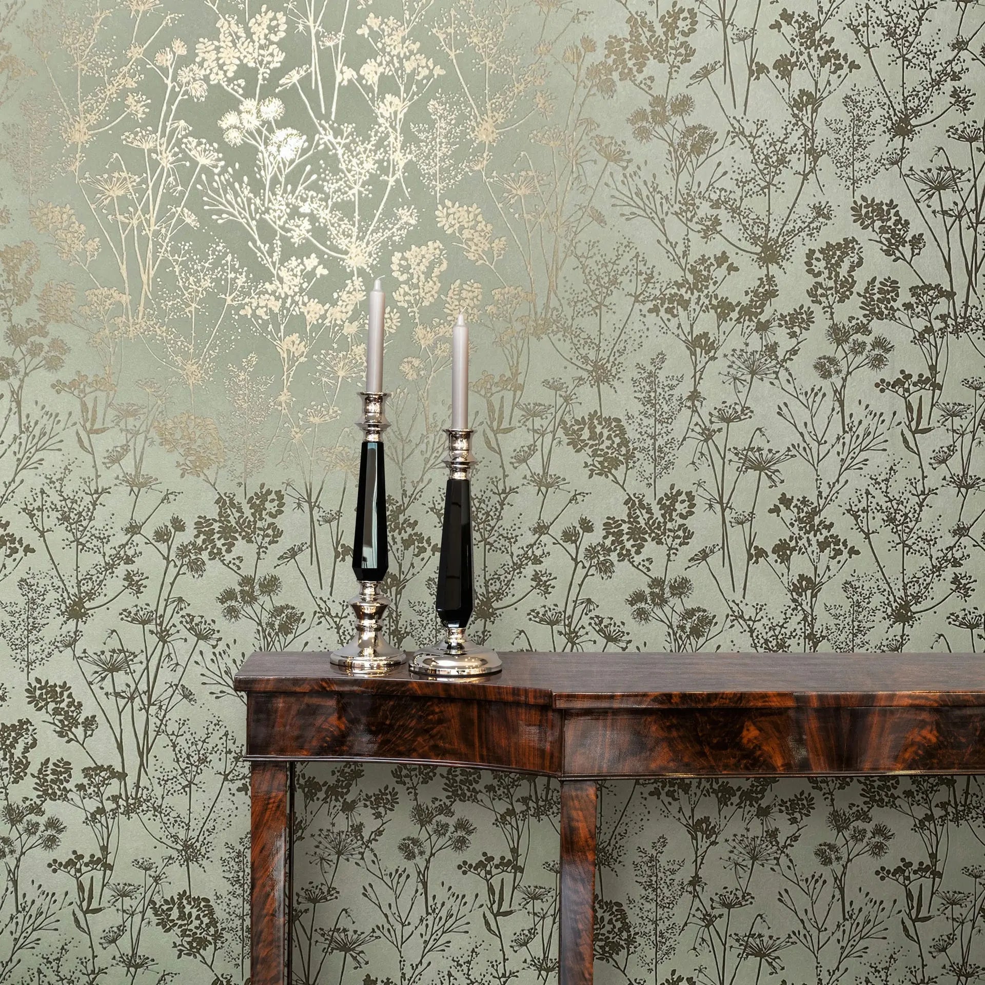 Marburg Mona 82465 Green wallpaper with metallic gold and dark green botanical silhouettes behind a dark wood console table with two tall candlesticks and white taper candles.