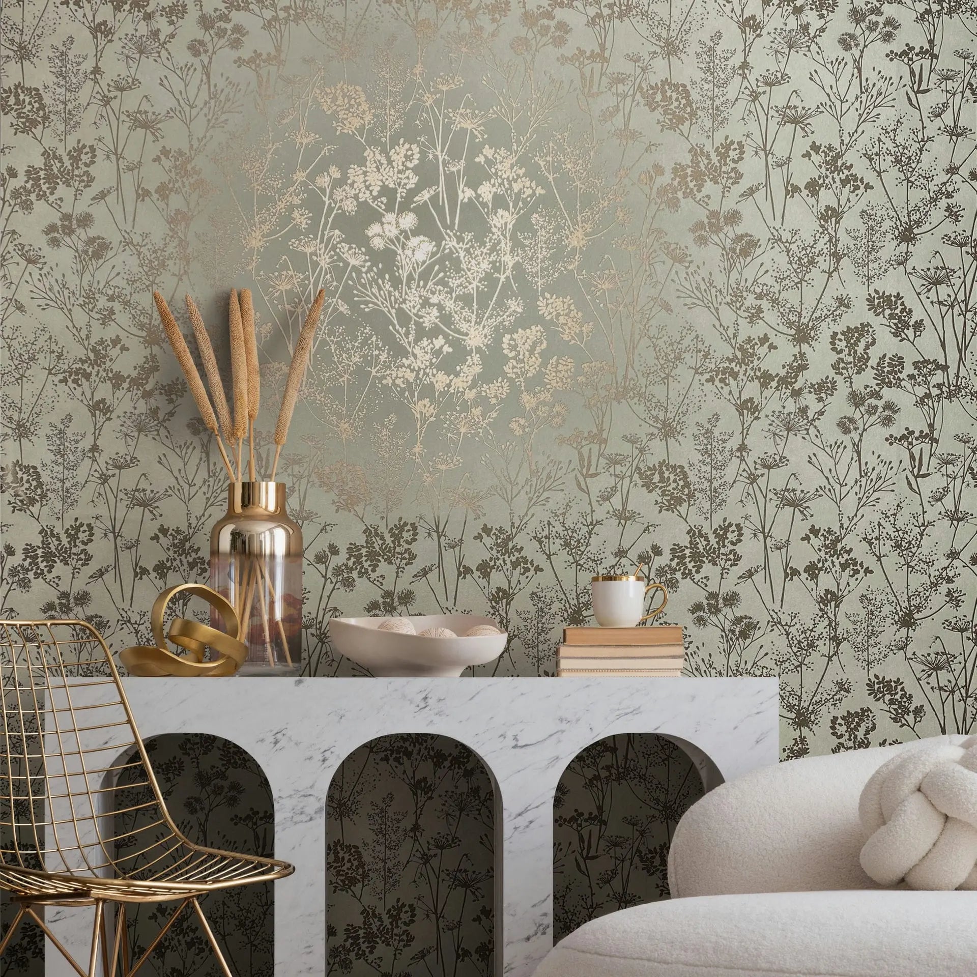Marburg Mona 82465 Green wallpaper with metallic gold botanical silhouettes shown in a styled living room scene with a marble console, glass vase with dried stems, decorative rings, shallow bowl, stack of books with a small cup, a gold wire chair, and a cream upholstered chair.
