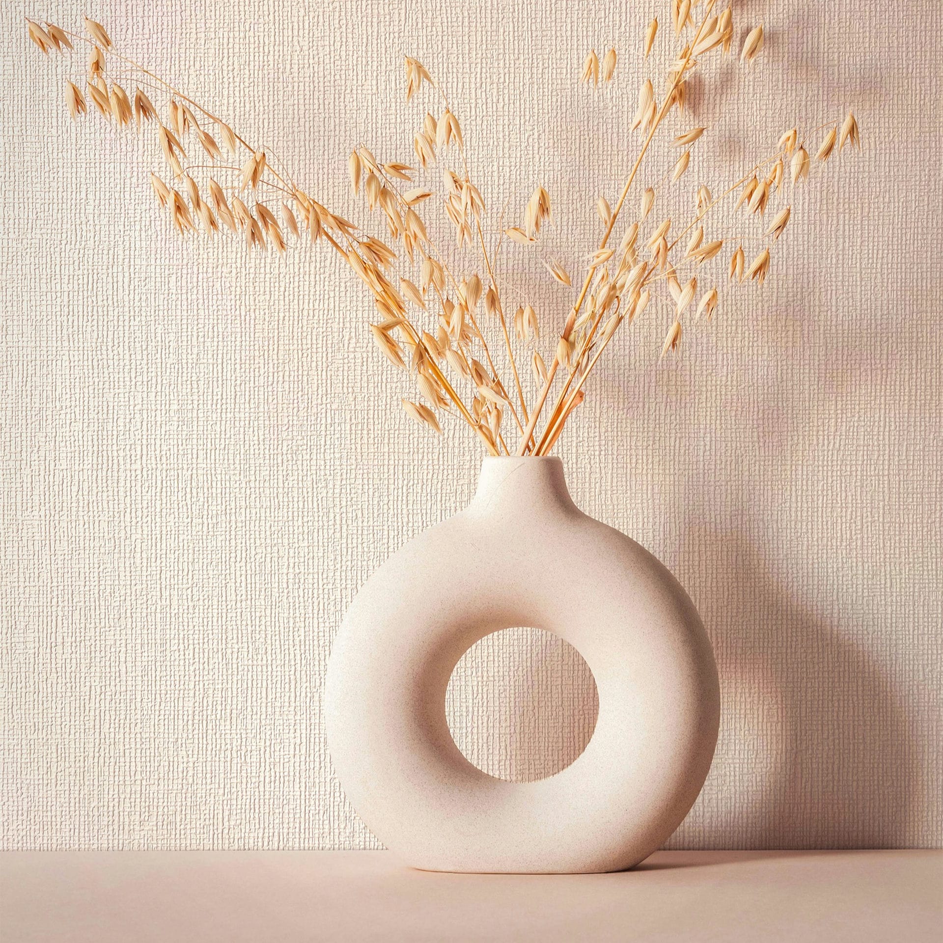 Marburg Mona 82466 Beige textured wallpaper with subtle irregular linear ridges shown behind a round ceramic donut‑shaped vase holding dried stems on a tabletop.