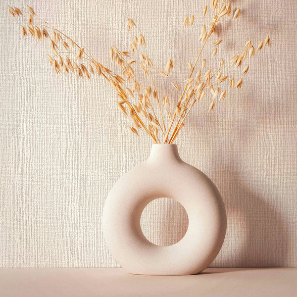 Marburg Mona 82466 Beige textured wallpaper with subtle irregular linear ridges shown behind a round ceramic donut‑shaped vase holding dried stems on a tabletop.