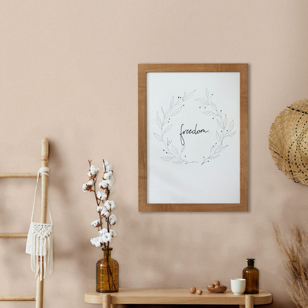 Marburg Mona 82466 Light beige textured wallpaper shown as a feature wall behind a framed print reading “freedom,” a wood console with an amber bottle holding cotton stems, a small cup and bottle, a leaning ladder with a macramé piece, and a straw hat on the wall.