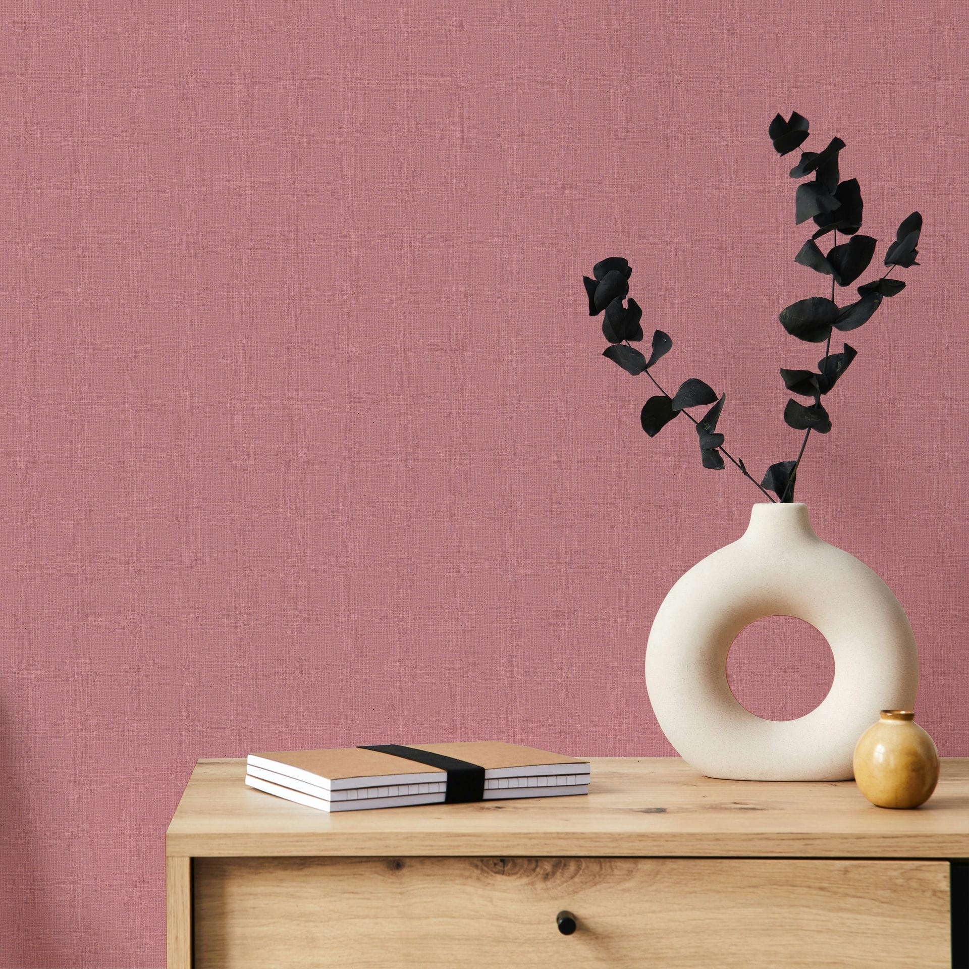 Marburg Mona 82467 Solid pink textured wallpaper behind a light wood cabinet topped with a round cutout ceramic vase holding dark foliage, a small round vase, and stacked notebooks.