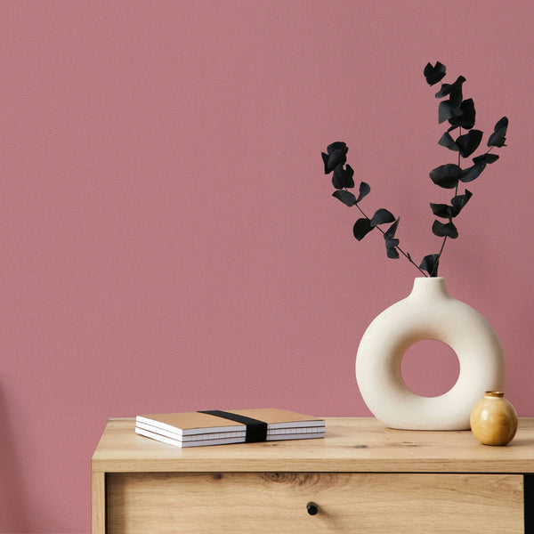 Marburg Mona 82467 Solid pink textured wallpaper behind a light wood cabinet topped with a round cutout ceramic vase holding dark foliage, a small round vase, and stacked notebooks.