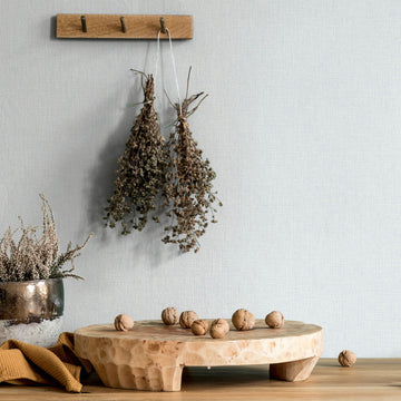 Marburg Mona 82473 Light neutral textured wallpaper with a fine crosshatch. A wooden peg rail holds two dried herb bundles above a carved wood riser on a tabletop with scattered walnuts and a reflective vase of dried stems at left.