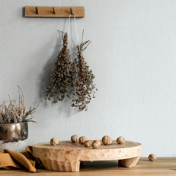 Marburg Mona 82473 Light neutral textured wallpaper with a fine crosshatch. A wooden peg rail holds two dried herb bundles above a carved wood riser on a tabletop with scattered walnuts and a reflective vase of dried stems at left.
