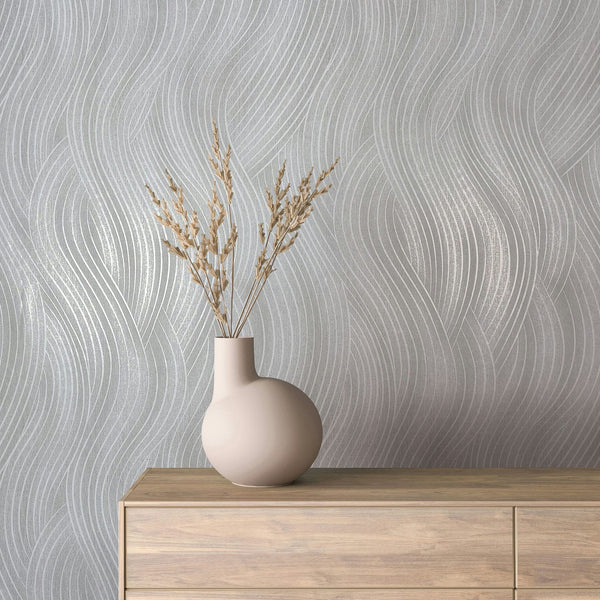 Marburg Mona 82474 Gray wallpaper with flowing wavy lines shown behind a light wood dresser with a round matte vase holding dried stems.