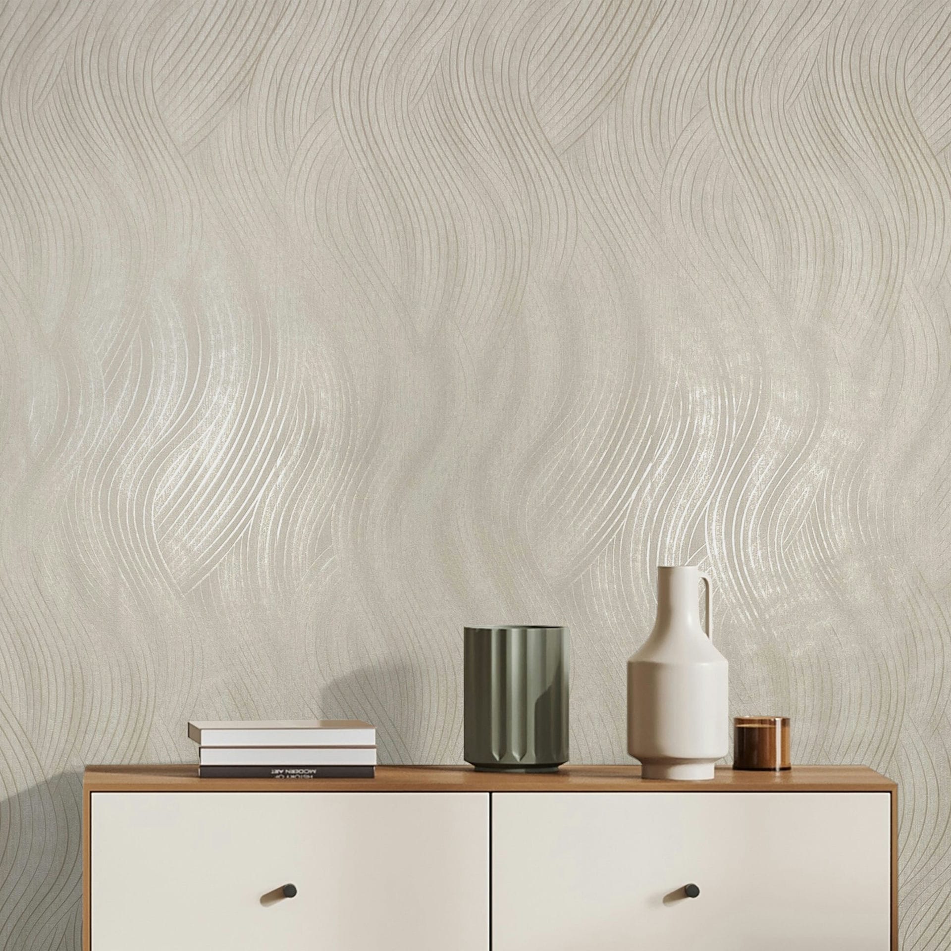 Marburg Mona 82475 Light beige wallpaper with a flowing wavy line pattern and subtle sheen, shown behind a wood‑top sideboard with stacked books, a green ribbed vase, a white handled vessel, and a small metallic cup.