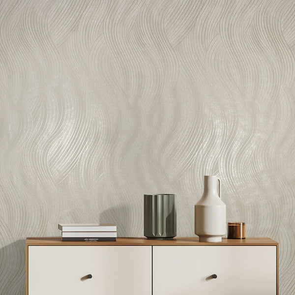 Marburg Mona 82475 Light beige wallpaper with a flowing wavy line pattern and subtle sheen, shown behind a wood‑top sideboard with stacked books, a green ribbed vase, a white handled vessel, and a small metallic cup.