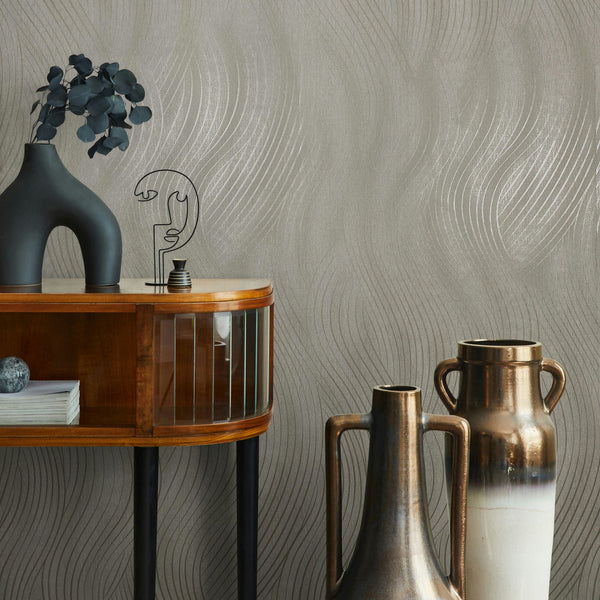 Marburg Mona 82475 Beige-gray wallpaper with shimmering wavy lines shown with a wood console, a black sculptural vase with foliage, a wire face ornament, and two tall metallic vases.
