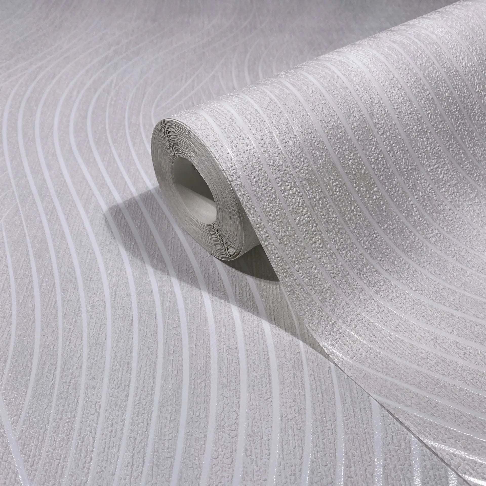 Marburg Mona 82476 Close-up of a white wallpaper roll with curved wavy stripes and textured bands, shown partially unrolled on a matching surface.