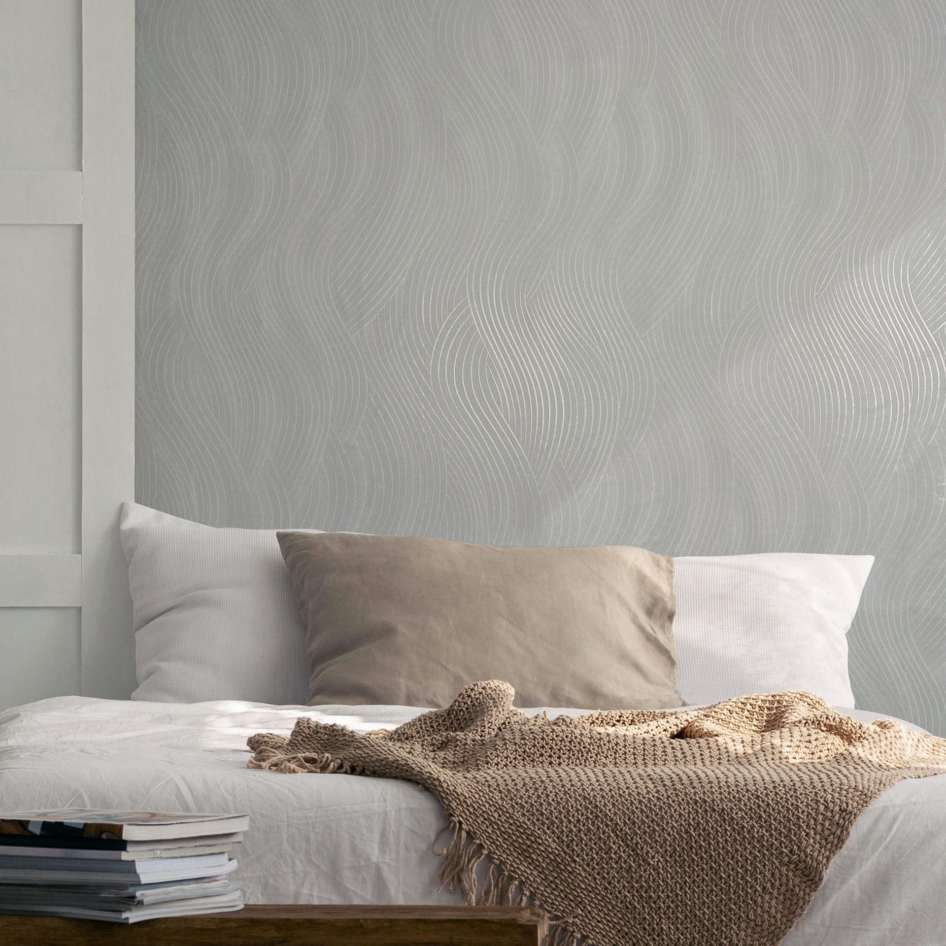 Marburg Mona 82476 Light gray wallpaper with a flowing wavy line pattern behind a bed with pillows and a knitted throw.