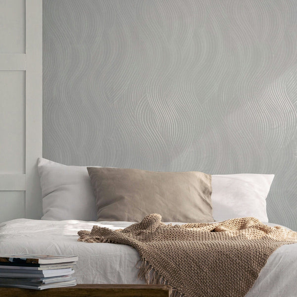 Marburg Mona 82476 Light gray wallpaper with a flowing wavy line pattern behind a bed with pillows and a knitted throw.