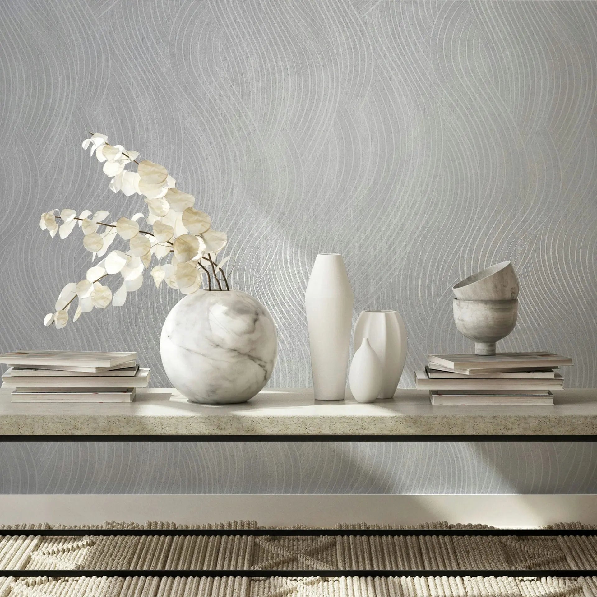 Marburg Mona 82476 Light grey wallpaper with subtle wavy lines behind a console shelf styled with a spherical stone-look vase holding delicate stems, white vases, a small stone bowl, and stacked books over a textured rug.