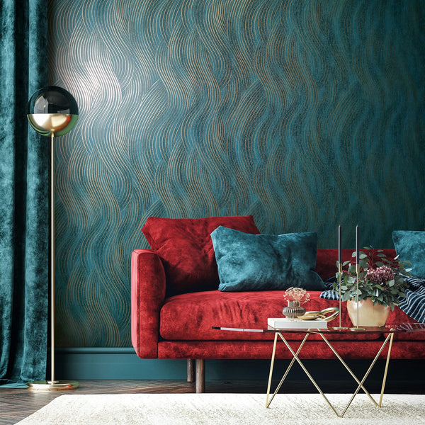 Marburg Mona 82477 Living room with wallpaper featuring metallic gold wavy lines on a deep teal background. A red sofa with teal pillows sits in front, with a gold floor lamp, teal curtain, and a glass coffee table holding a potted plant and candles on a light rug.