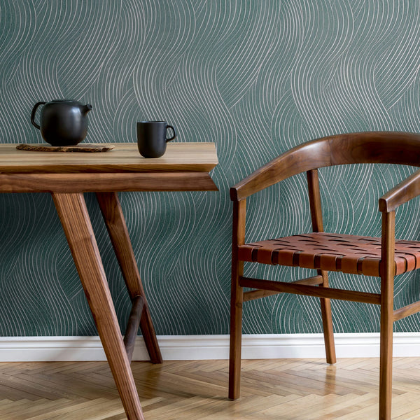 Marburg Mona 82478 Green wallpaper with sinuous silver lines behind a wooden table with a black teapot and mug and a wooden chair on a herringbone floor.
