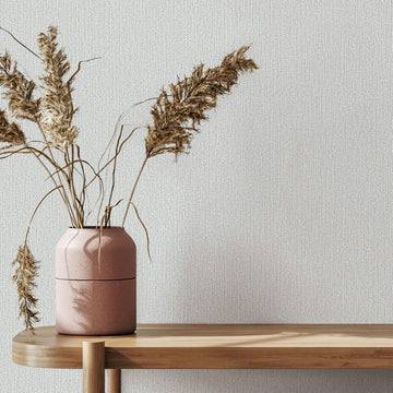 Marburg Mona 82479 Light gray textured wallpaper with fine vertical lines behind a wooden console topped with a pink vase holding dried grasses.