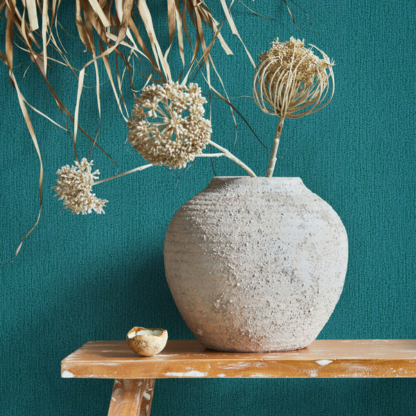Marburg Mona 82482 Textured green wallpaper with a subtle vertical pattern, shown behind a large rough ceramic vase on a wooden bench with dried botanical stems.