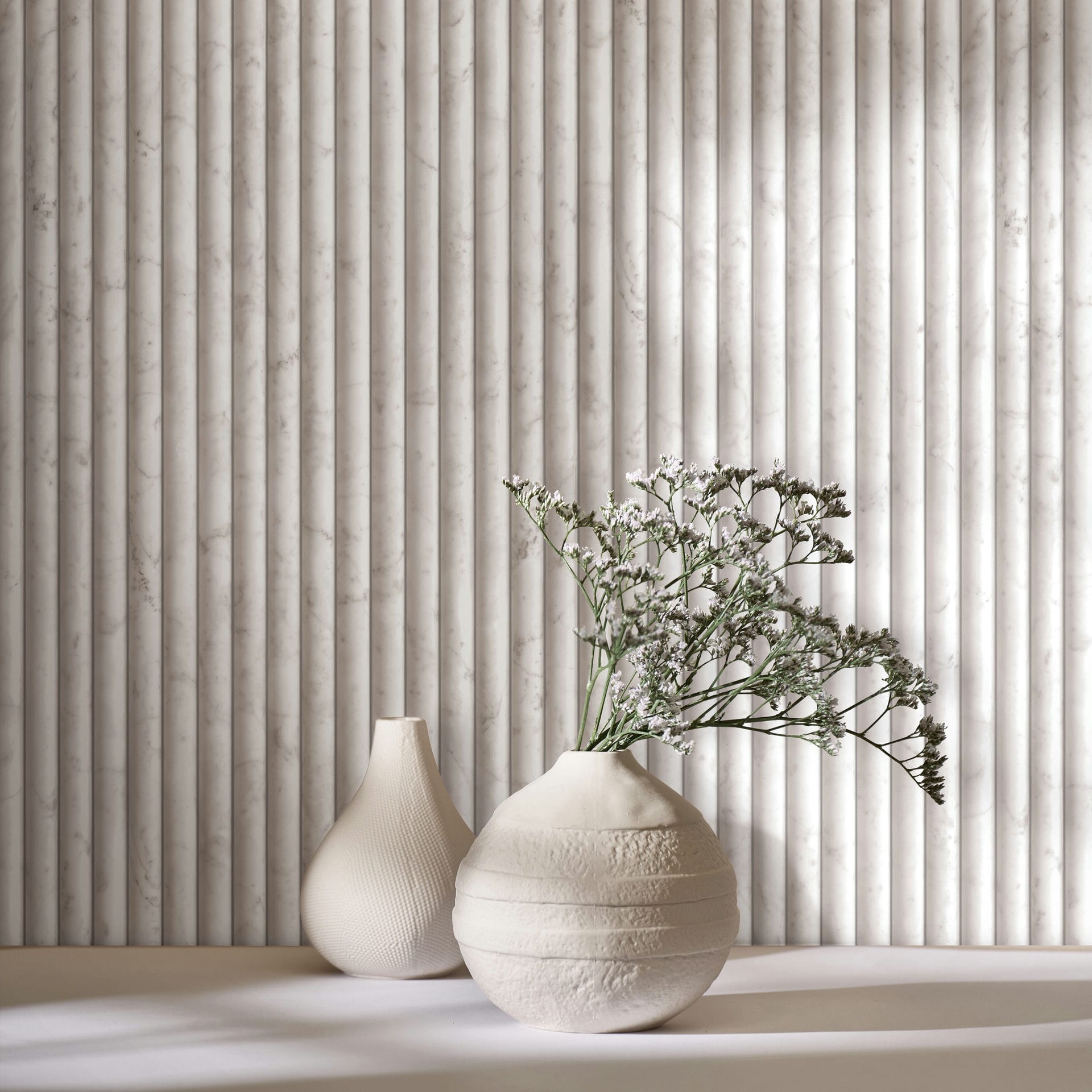 Marburg Mona 89004 Close-up of light beige-gray wallpaper printed with a vertical fluted marble effect, with two cream ceramic vases on a surface, one holding small white flowers.