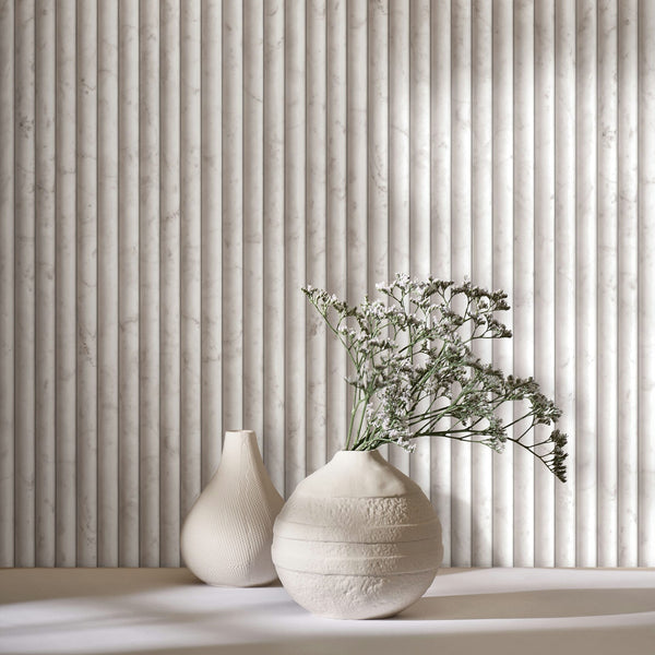 Marburg Mona 89004 Close-up of light beige-gray wallpaper printed with a vertical fluted marble effect, with two cream ceramic vases on a surface, one holding small white flowers.