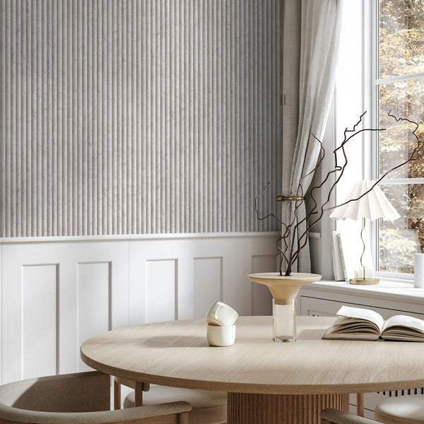 Marburg Mona 89004 Light beige-gray wallpaper with narrow vertical ribbed stripes above white wainscoting, shown in a dining area with a round table, chairs, a window with curtains, a table lamp, an open book, a cup, and a vase with branches.