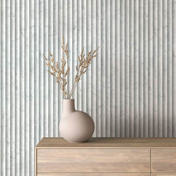 Marburg Mona 89005 Light gray marble-look wallpaper with narrow vertical fluted stripes, shown behind a beige vase with dried stems on a wood dresser.