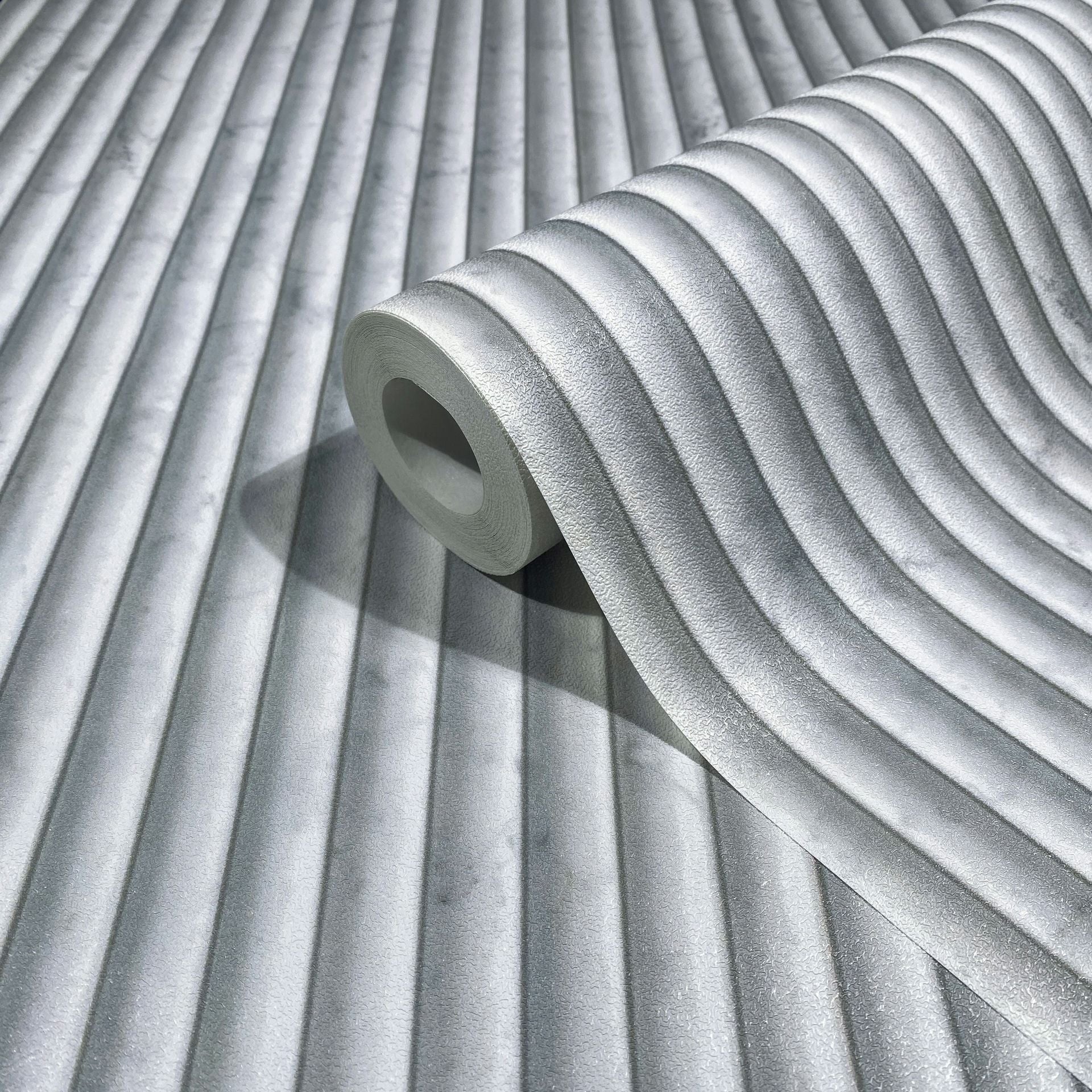 Marburg Mona 89005 A gray wallpaper roll with softly curved vertical ribs, shown resting on the same patterned wallpaper.