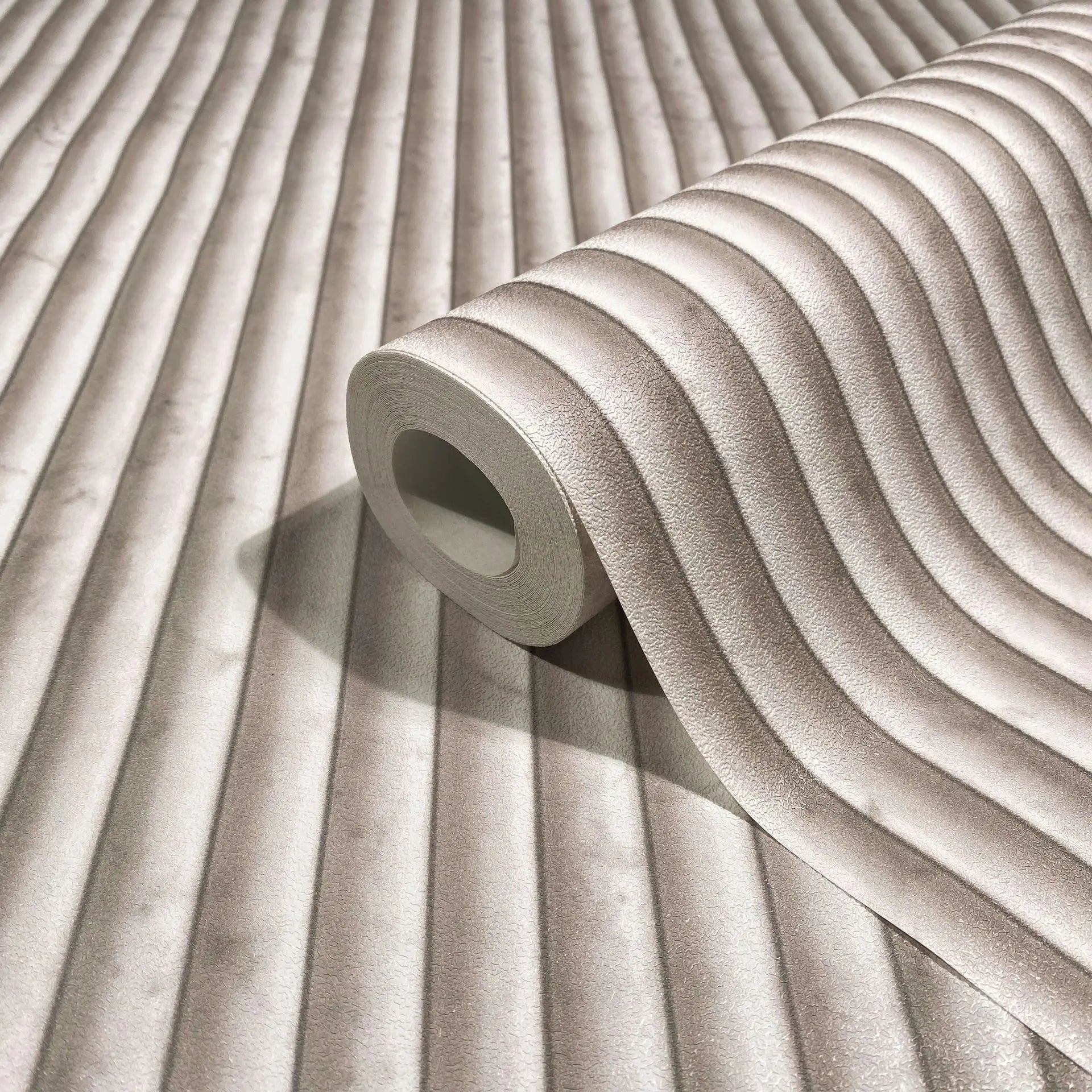 Marburg Mona 89006 Wallpaper roll on a matching surface showing a beige ribbed, softly wavy stripe pattern with a subtle 3D effect.