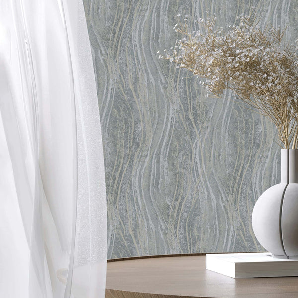 Marburg Naomi 82494 Styled detail showing gray wallpaper with flowing vertical wavy lines and a textured look. In front are a sheer curtain, a round vase with dried stems, and a book on a round wooden table.