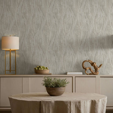 Marburg Naomi 82495 Beige and gray wallpaper with vertical wavy lines behind a light wood sideboard, a modern table lamp, stacked books, a wooden sculpture, a bowl of green fruit, and a round table with a potted plant.