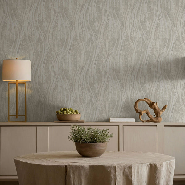 Marburg Naomi 82495 Beige and gray wallpaper with vertical wavy lines behind a light wood sideboard, a modern table lamp, stacked books, a wooden sculpture, a bowl of green fruit, and a round table with a potted plant.