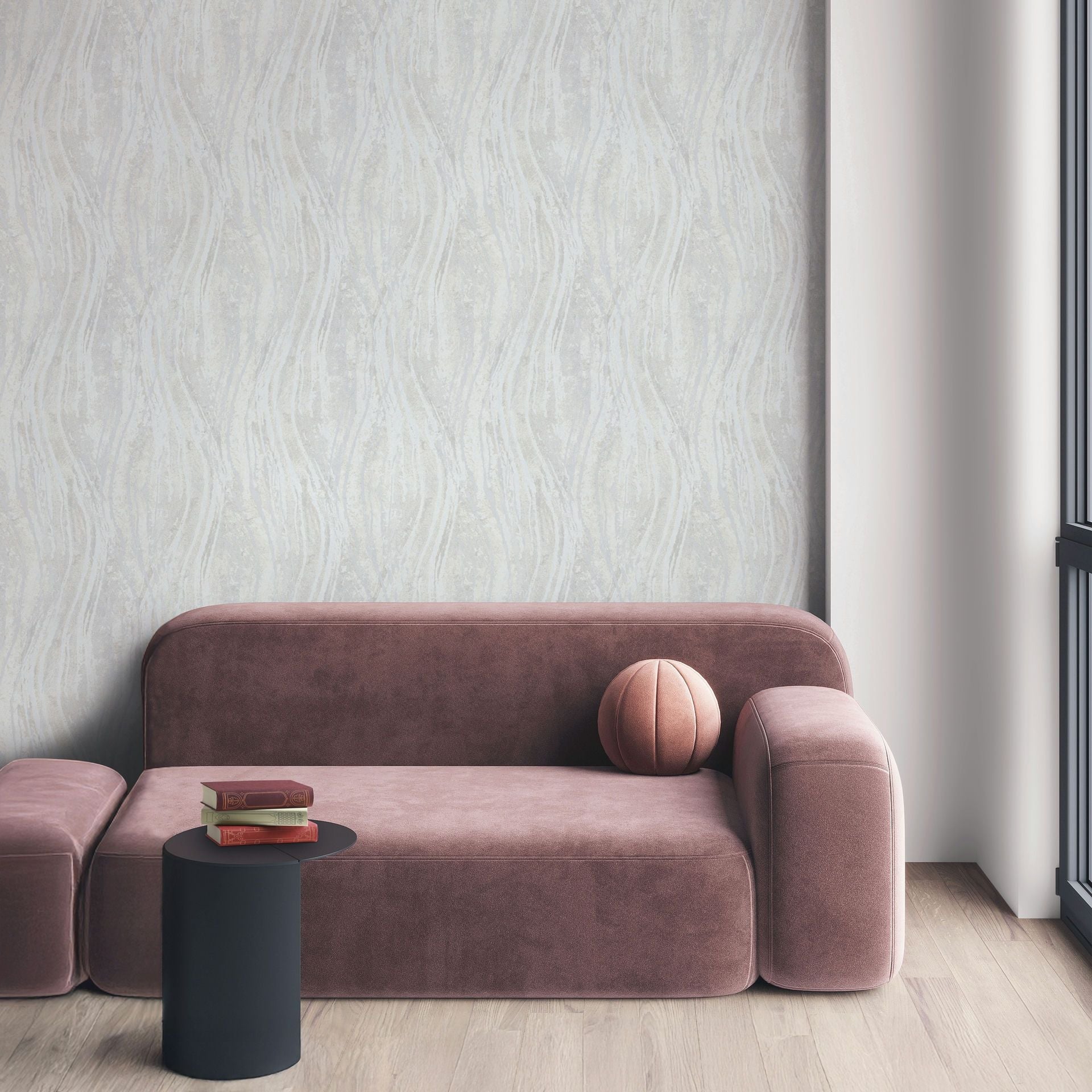 Marburg Naomi 82497 Light gray wallpaper with subtle vertical wavy stripes behind a modern sofa. A small black side table with stacked books, a round cushion, an ottoman, wood flooring, and a window frame are visible.