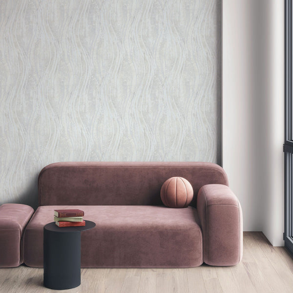 Marburg Naomi 82497 Light gray wallpaper with subtle vertical wavy stripes behind a modern sofa. A small black side table with stacked books, a round cushion, an ottoman, wood flooring, and a window frame are visible.