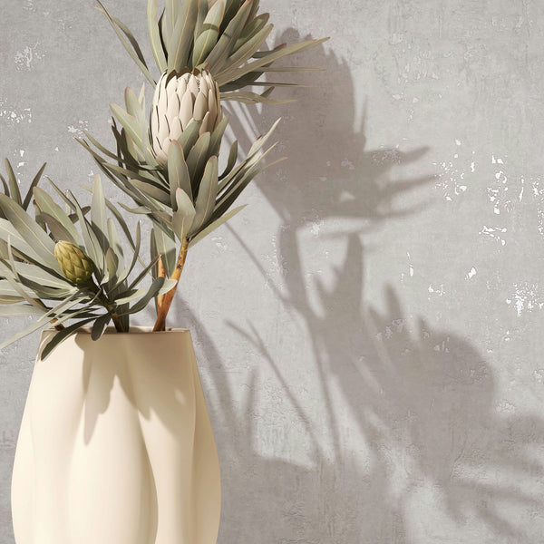 Marburg Naomi 82499 Close-up of light gray textured wallpaper with a subtle plaster effect, shown behind a cream ceramic vase holding protea stems and casting soft shadows.