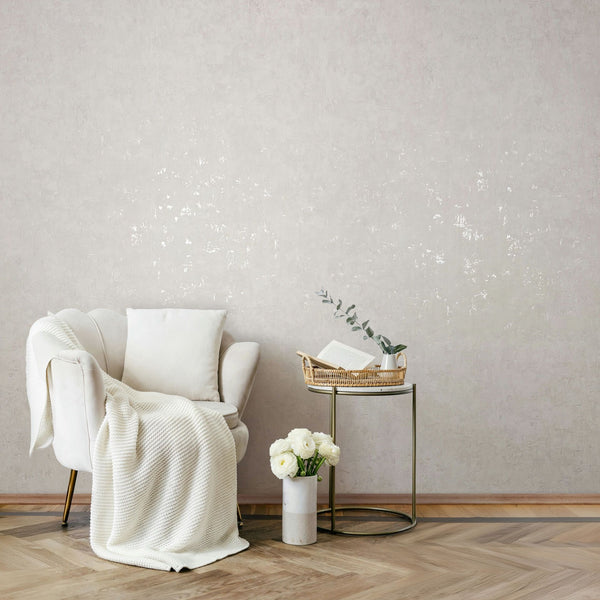 Marburg Naomi 82499 Light textured wallpaper with subtle reflective accents behind an upholstered armchair with a knit throw, a round side table holding a basket, book, and small vase with greenery, and a floor vase of flowers on parquet flooring.