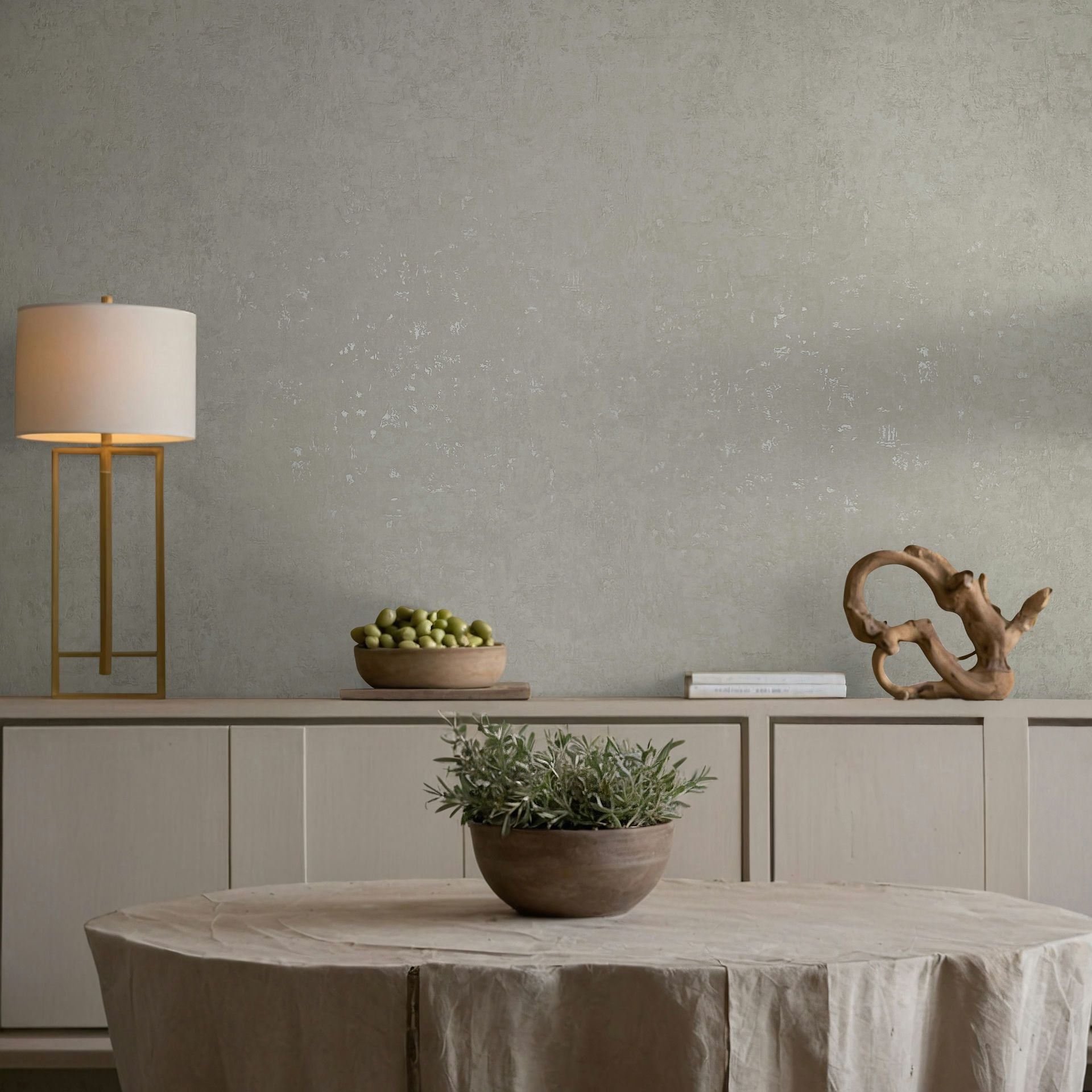 Marburg Naomi 82500 Gray textured wallpaper with a subtle mottled pattern behind a light wood sideboard holding a lamp, a bowl of green fruit, books, and a sculptural wood object, with a round table in front topped with a potted plant.