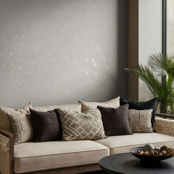 Marburg Naomi 82500 Gray textured wallpaper with a subtle distressed effect behind a light sofa with assorted pillows, a round coffee table with a decorative bowl, and a potted palm by a window.