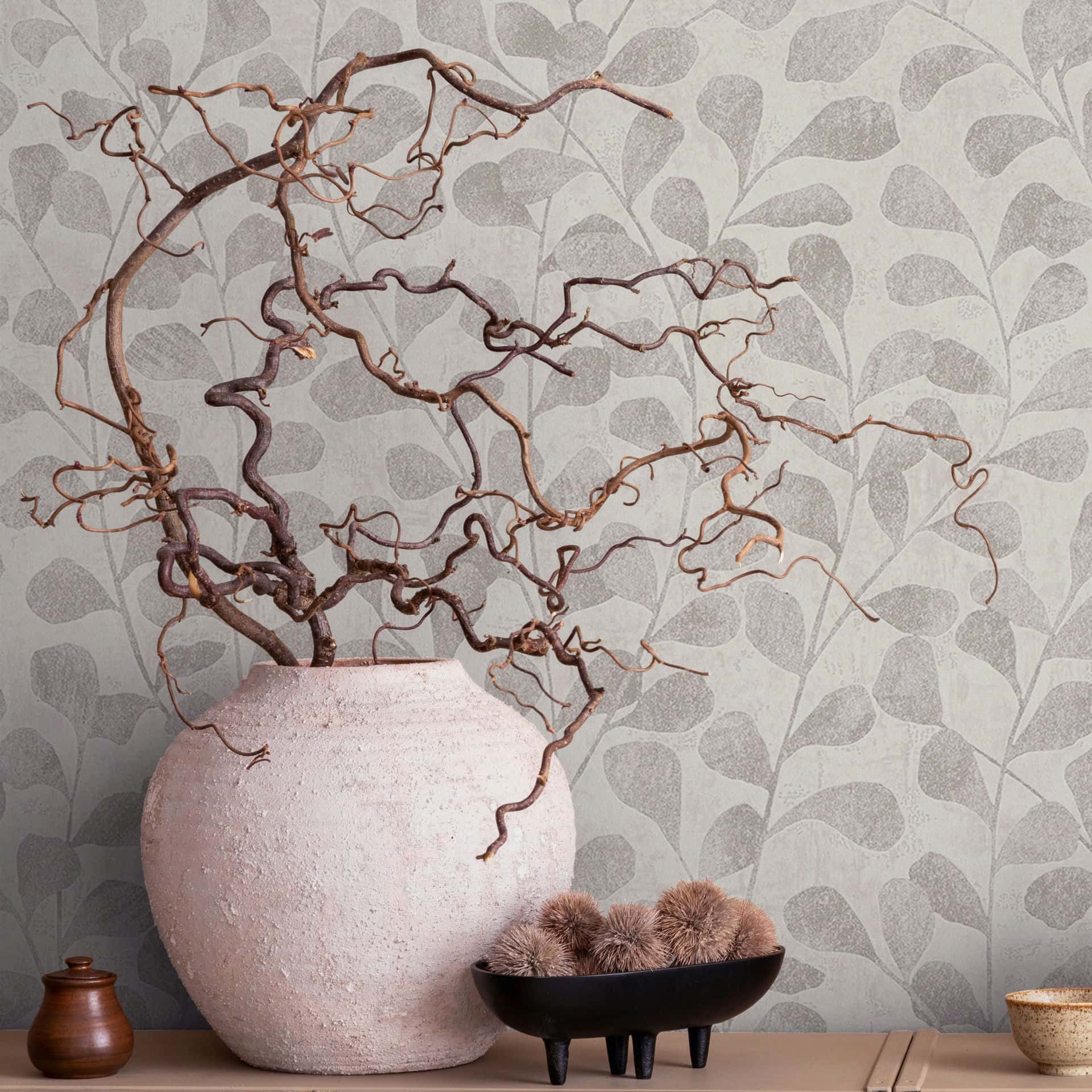 Marburg Naomi 82502 Botanical wallpaper with overlapping leaf silhouettes in gray-beige tones, shown behind a large pale textured vase filled with twisted branches, a small wooden lidded jar, a black footed bowl holding fuzzy decorative balls, and a small speckled cup on a low-s.