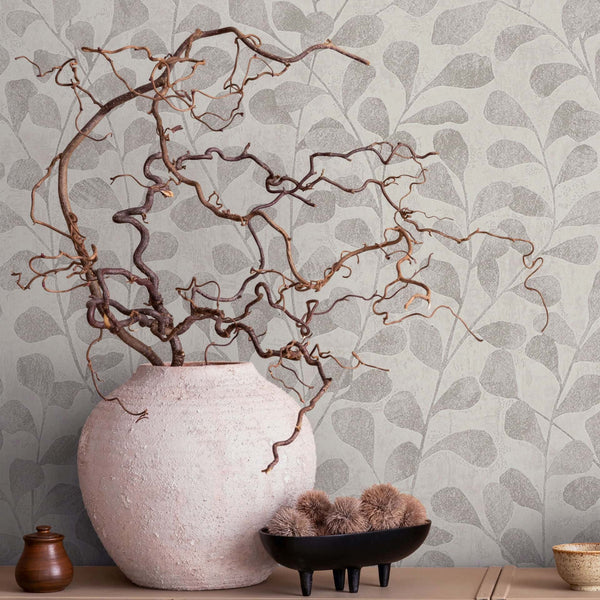 Marburg Naomi 82502 Botanical wallpaper with overlapping leaf silhouettes in gray-beige tones, shown behind a large pale textured vase filled with twisted branches, a small wooden lidded jar, a black footed bowl holding fuzzy decorative balls, and a small speckled cup on a low-s.