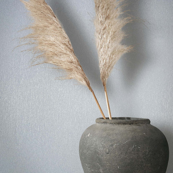Marburg Naomi 82535 Close-up of light blue textured wallpaper with a subtle vertical shimmer, styled with a gray stone vase holding two pampas grass stems.