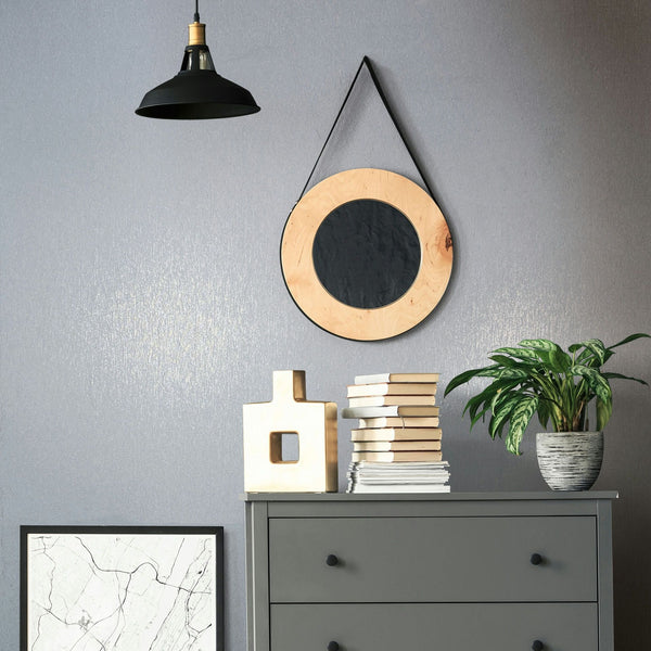 Marburg Naomi 82535 Blue-gray textured wallpaper with a soft metallic sheen shown behind a round hanging mirror, black pendant light, gray dresser with stacked books and a decorative object, a potted plant, and a framed print.