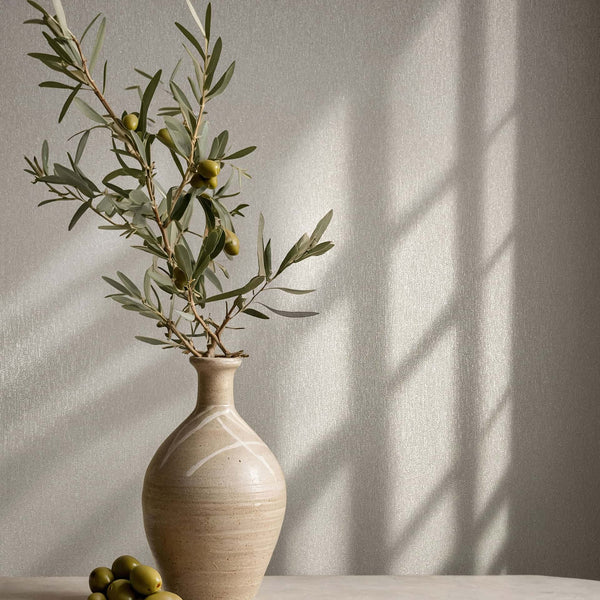 Marburg Naomi 82536 Beige-gray textured wallpaper shown behind a ceramic vase holding olive branches, with windowpane shadows cast across the wall.