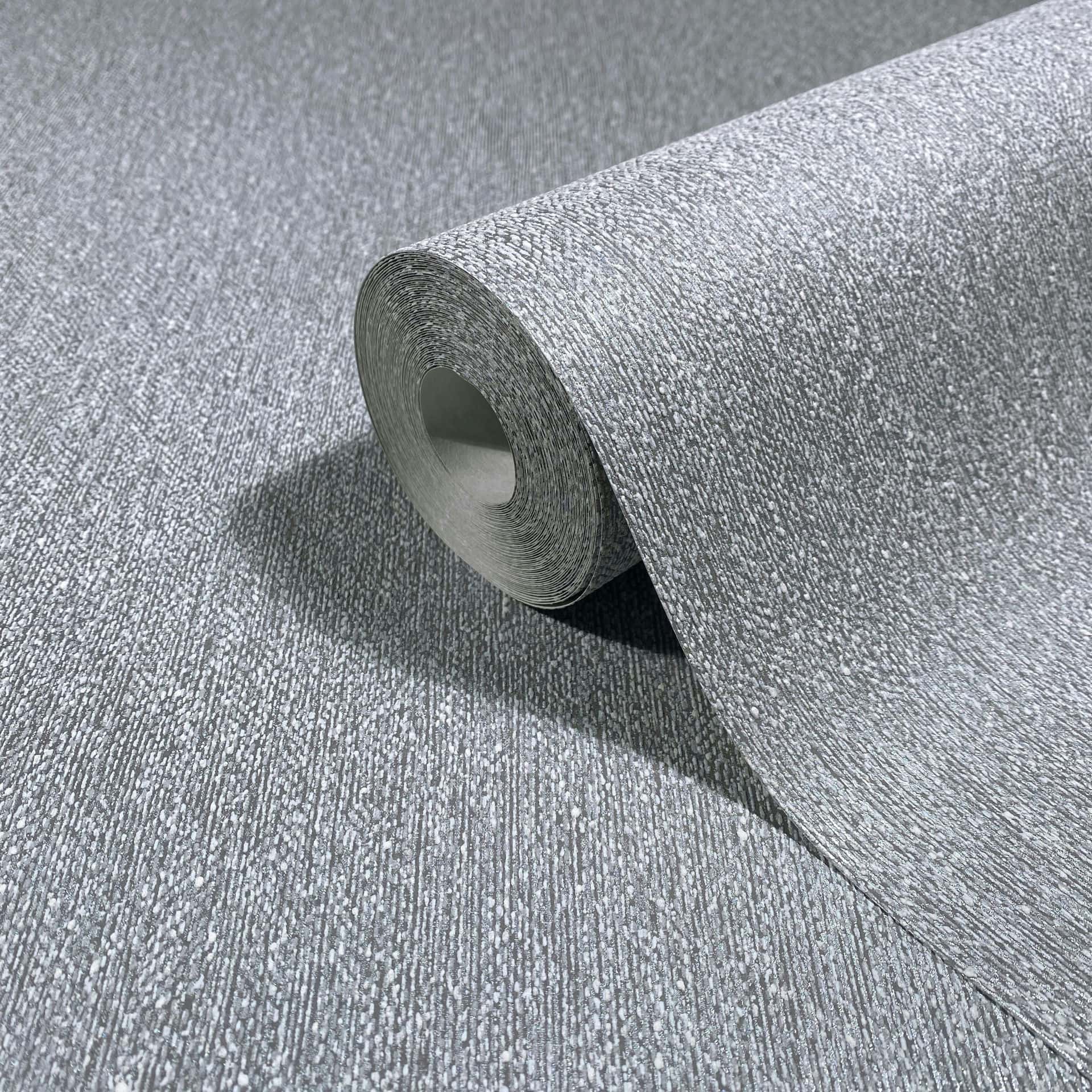 Marburg Naomi 82537 Close-up of a gray textured wallpaper roll, partly unrolled on a matching surface with fine vertical striations and speckled detail.
