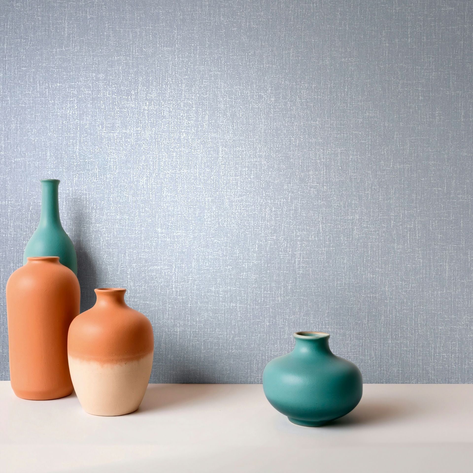Marburg Naomi 82542 Light blue wallpaper with subtle crosshatch texture, shown behind four ceramic vases in teal and terracotta on a white surface.