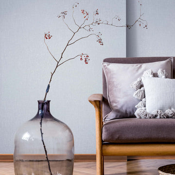 Marburg Naomi 82542 Light grey textured wallpaper behind a large glass floor vase holding a bare branch with red berries next to a cushioned wooden armchair.