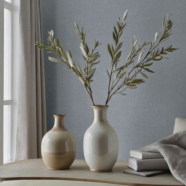 Marburg Naomi 82543 Light gray textured wallpaper with fine vertical striations behind a tabletop scene featuring two ceramic vases, one with leafy branches, stacked books, a draped fabric, and a window with a curtain.