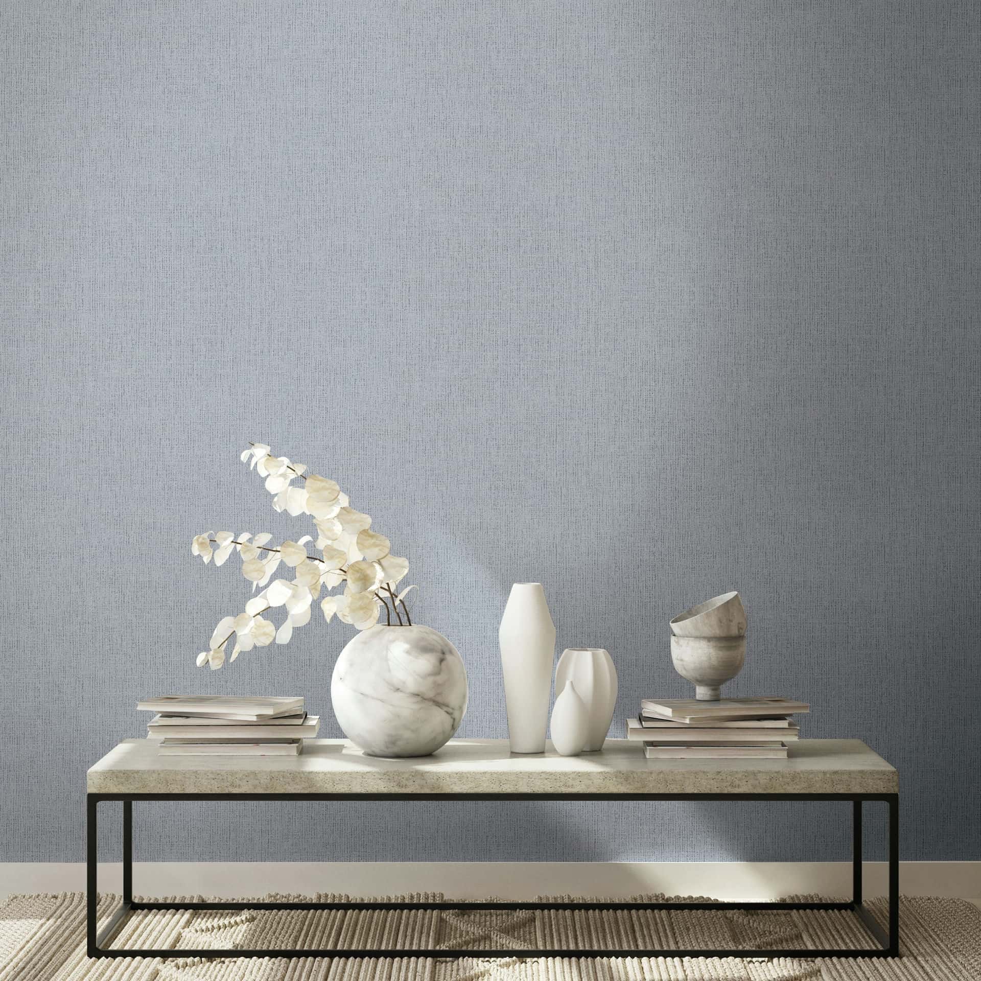 Marburg Naomi 82543 Gray textured wallpaper with a fine woven look behind a low table styled with a marble vase holding dried stems, two white vases, a marble bowl, and stacked books on a rug.