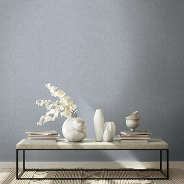 Marburg Naomi 82543 Gray textured wallpaper with a fine woven look behind a low table styled with a marble vase holding dried stems, two white vases, a marble bowl, and stacked books on a rug.