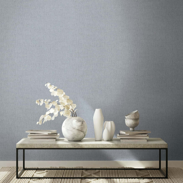 Marburg Naomi 82543 Gray textured wallpaper with a fine woven look behind a low table styled with a marble vase holding dried stems, two white vases, a marble bowl, and stacked books on a rug.