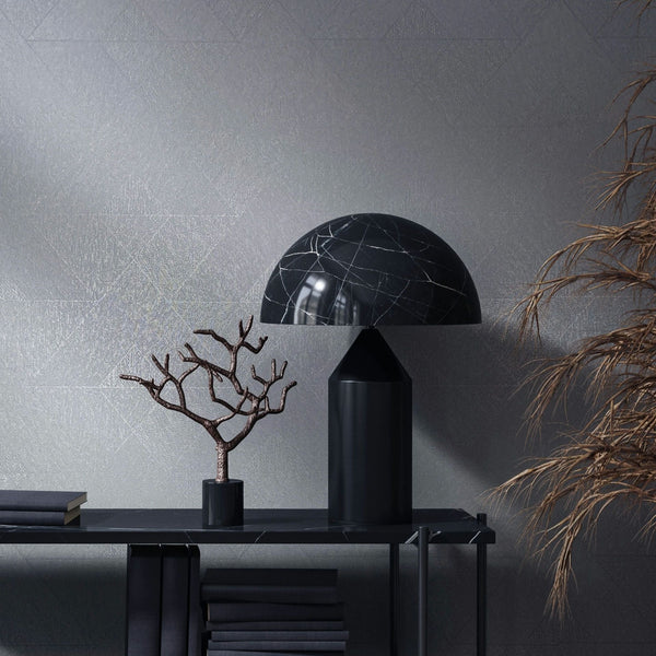 Marburg Naomi 82547 Tonal geometric wallpaper with a subtle line pattern shown behind a black dome table lamp on a slim console with stacked books and a small sculptural branch; dried foliage appears at the right edge.